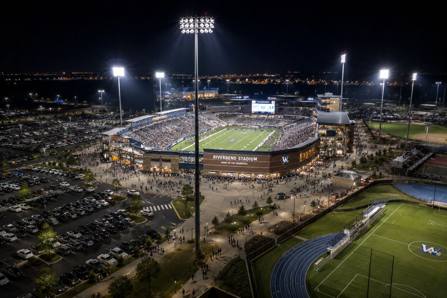 LED high mast lighting illuminating a large sports stadium with surrounding parking lots, pedestrian walkways, athletic fields, and nighttime event operations.